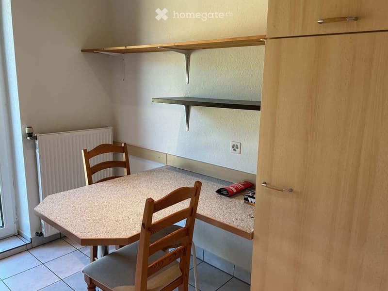 A small, furnished room with a round table, two wooden chairs, built-in shelves, and a radiator. The room has tiled flooring and light-colored walls.