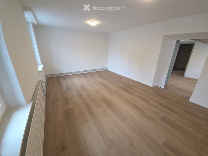 Empty room with light wooden flooring, white walls, and a radiator on the left side. The room has large windows and a visible doorway leading to another space.