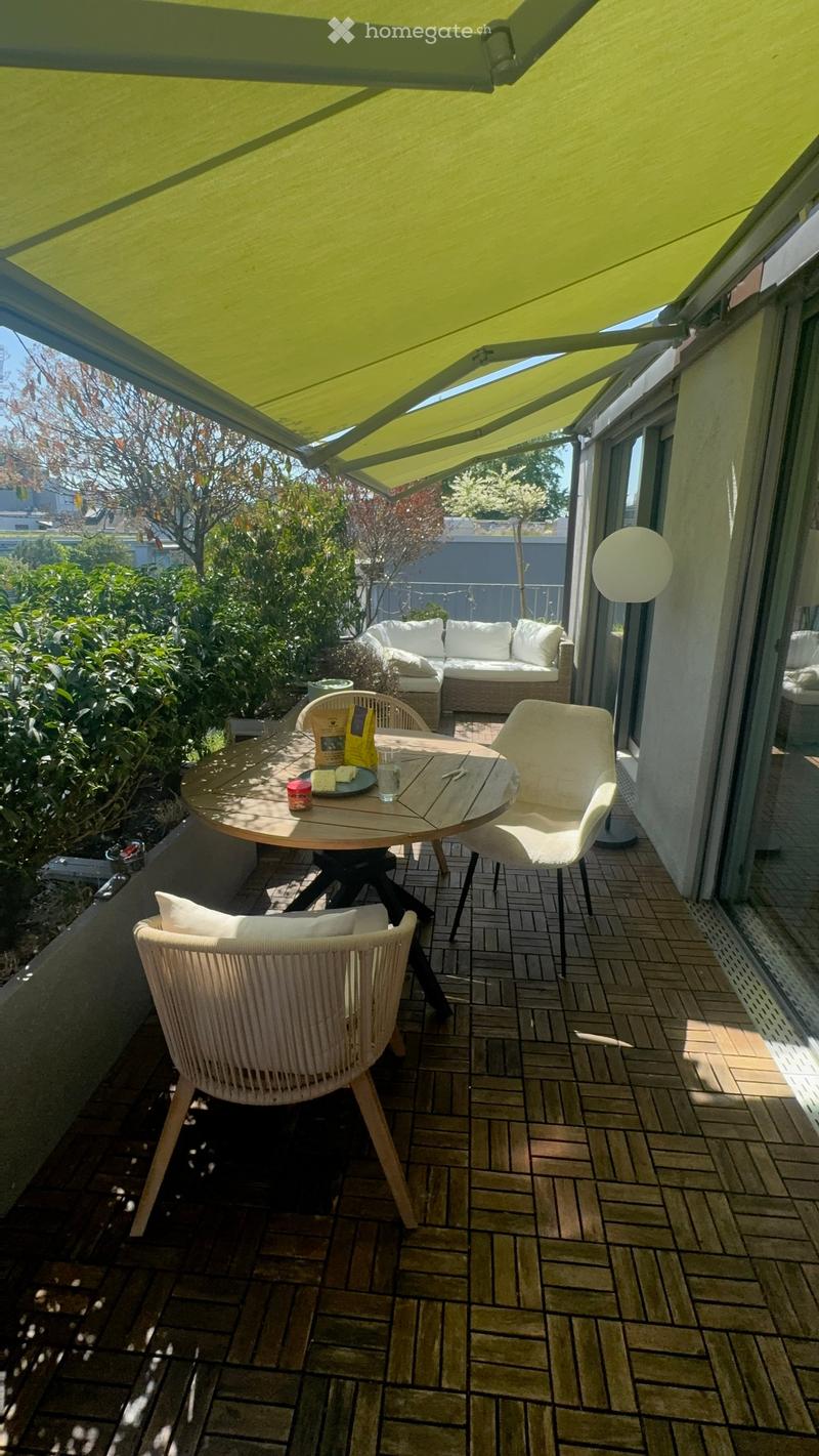 Balcony with a round wooden table, two wicker chairs, and a sofa in the background, covered by a green awning.