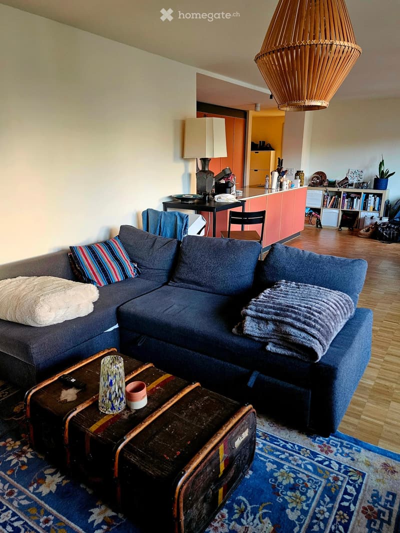 A modern living room with a blue sectional sofa, a wooden coffee table, a large hanging lamp, and a small kitchen area visible in the background.