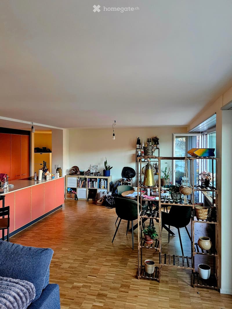 A modern, open-concept living and dining area with wooden flooring, a blue sofa, a dining table with chairs, shelves filled with books and decorative items, and a kitchen area with orange cabinets.