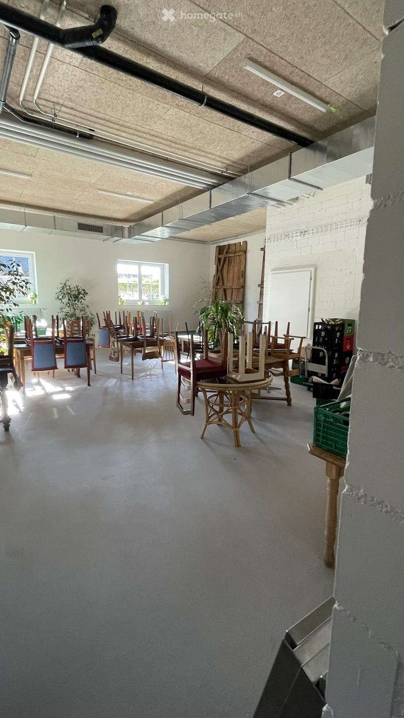 A spacious room with several wooden chairs and tables arranged, along with potted plants near the windows, and exposed ceiling beams.
