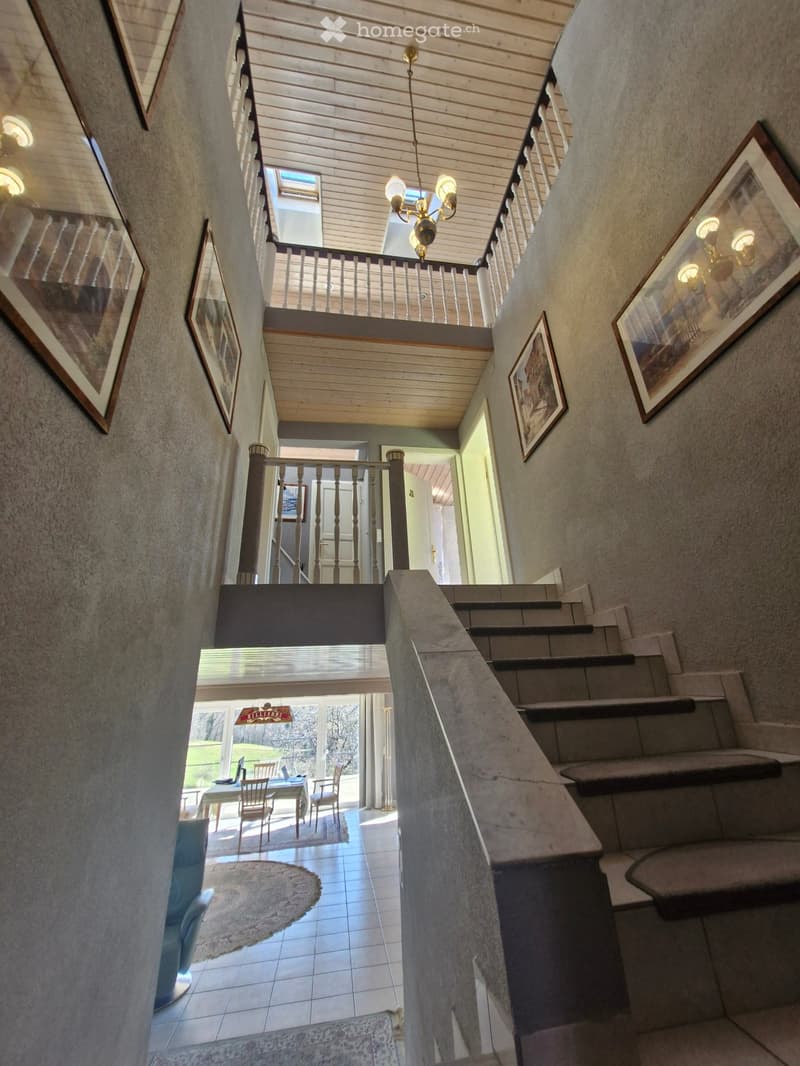Staircase with wooden handrail, tiled floor, wooden ceiling, chandelier, framed pictures on the wall, dining table with chairs, rug, balcony with railings, glass windows, view of the outside
