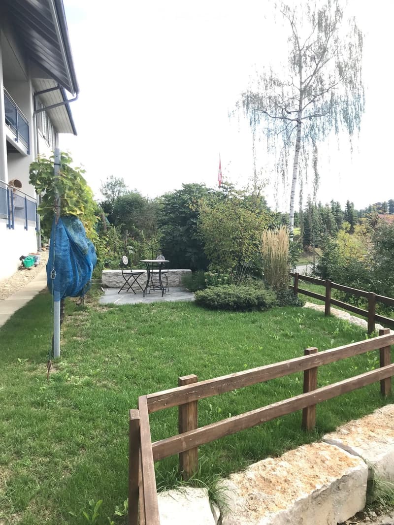 Garden with green grass, wooden fence, small patio area with a table and chairs, surrounded by trees and shrubs.