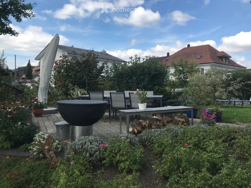 Well-maintained garden with a patio area, outdoor dining set, fire pit, potted plants, and neatly arranged shrubs and flowers.