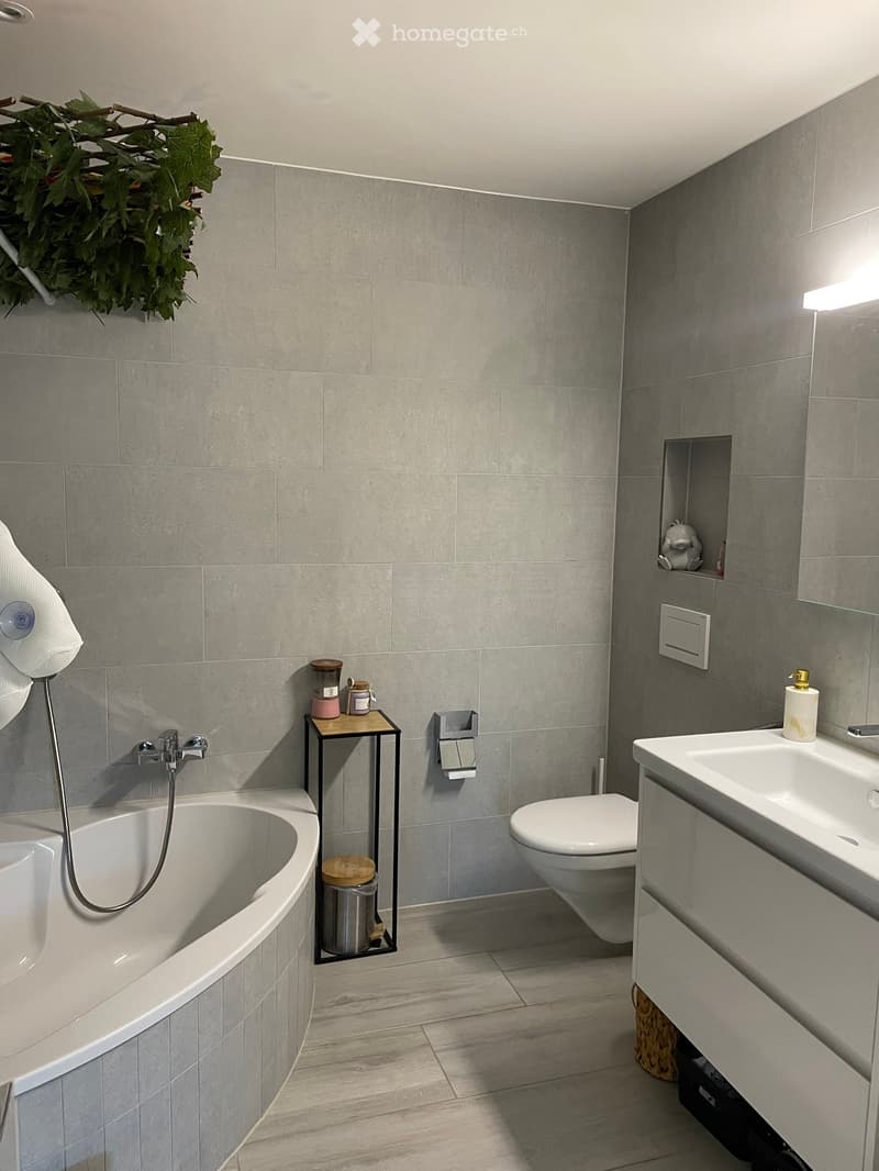 Modern bathroom with grey tiled walls, a bathtub, toilet, sink, and a small black table holding toiletries. A decorative leafy wreath is mounted on the wall above the bathtub.