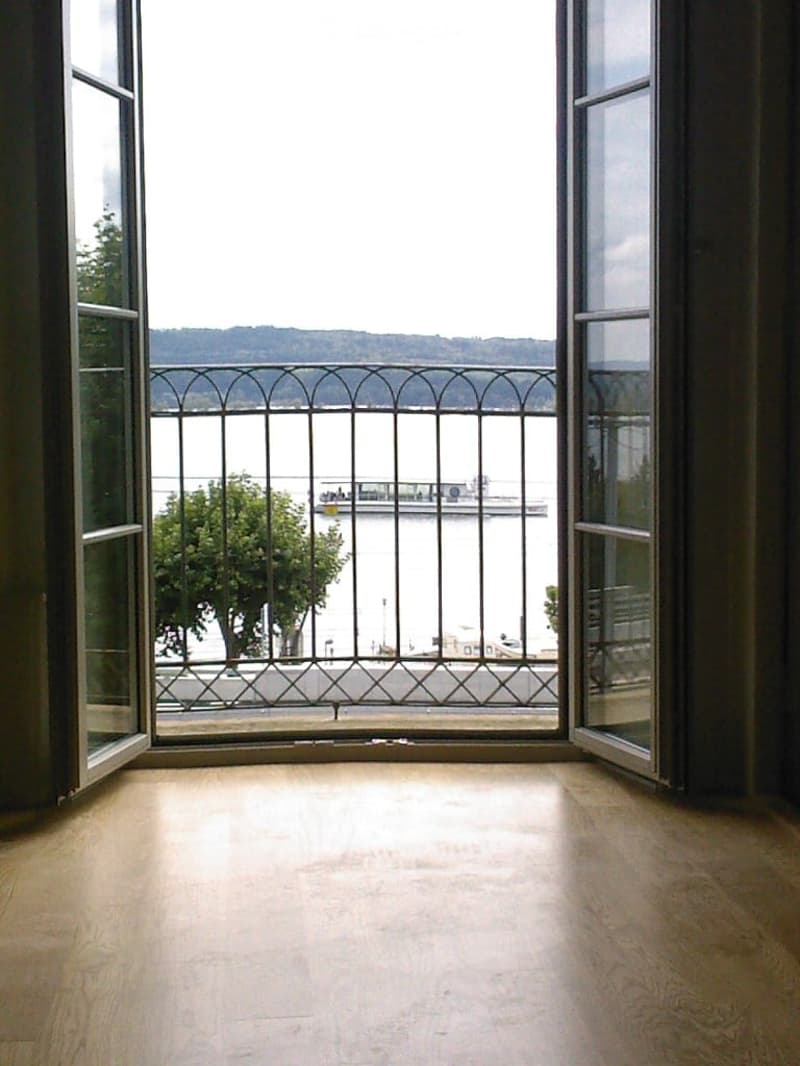 Empty room with wooden flooring and large windows opening to a balcony overlooking a scenic waterfront view.