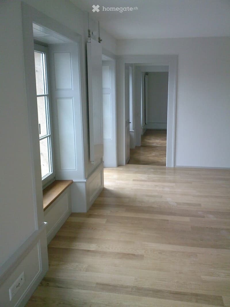 Empty hallway with light wooden flooring, white walls, and a bench seat under a window on the left side.