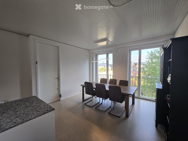 Bright dining area with a wooden table and six chairs, large windows and sliding doors leading to a balcony, modern lighting fixtures, and a dark cabinet.