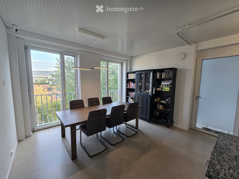Bright room with a wooden dining table and six chairs, a large bookshelf, and a cabinet filled with books and decorative items. Large windows and sliding doors lead to a balcony with a view of greenery.