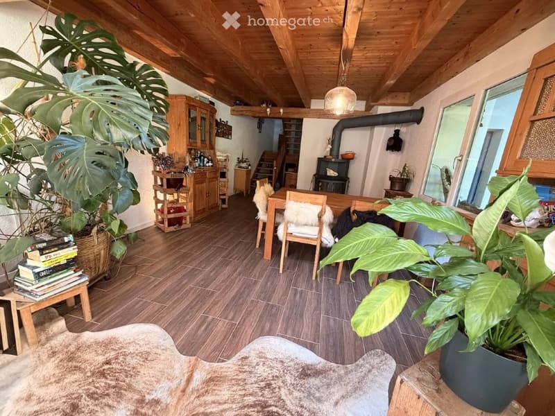 Cozy living and dining area with wooden beams, a dining table with chairs, a cabinet, a fireplace, and large potted plants.