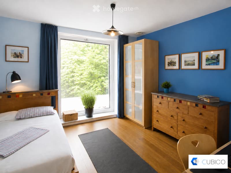 Bright bedroom with wooden bed, nightstand, chair, cabinet, and a large window with a view of greenery. Features a blue accent wall, framed pictures, and a potted plant on the windowsill.