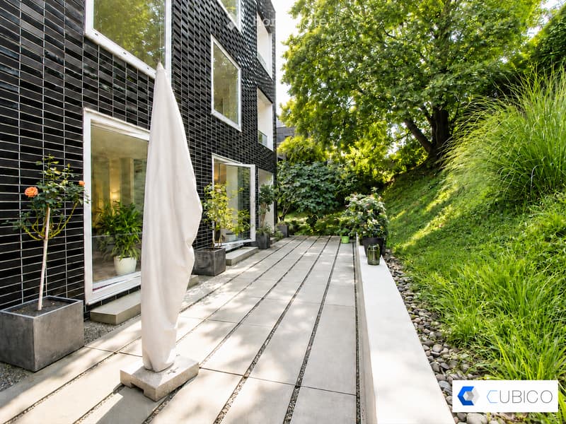 Modern outdoor patio area with a sleek, black-tiled house, large windows, and a well-maintained garden with various plants and trees. Features include a tall white parasol, stone pathway, and decorative planters.