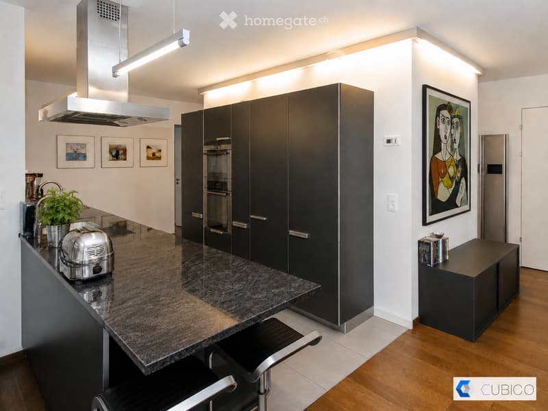 Modern kitchen with dark cabinets, marble countertop, stainless steel appliances, and a small seating area with a stool.