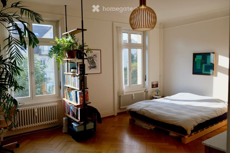 Bright bedroom with wooden flooring, large windows, a bed with white bedding, a tall bookshelf filled with books, a pendant light, and a small table with a chair near the window.