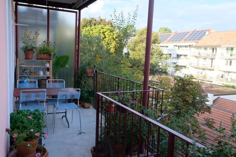 Balcony with a small table and chairs, surrounded by potted plants and a shelf with additional plants and items. The balcony overlooks a green area with trees and neighboring buildings.