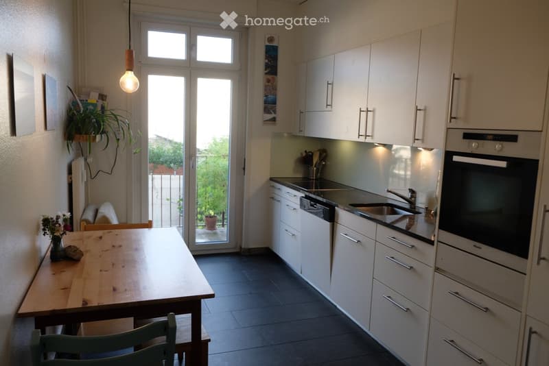 Modern kitchen with white cabinets, stainless steel appliances, a wooden dining table, and a hanging light fixture. A sliding glass door leads to a balcony with outdoor plants.