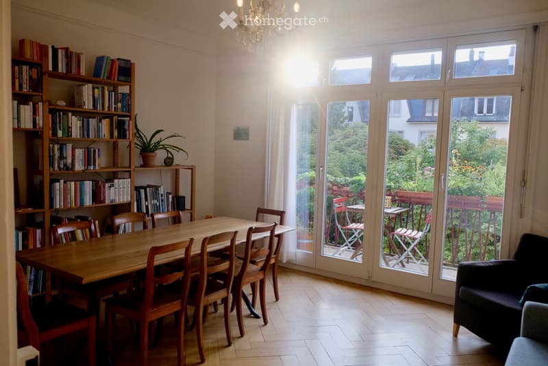 Bright dining area with a wooden table and chairs, a bookshelf filled with books, a potted plant, and a sofa near large windows opening to a garden.