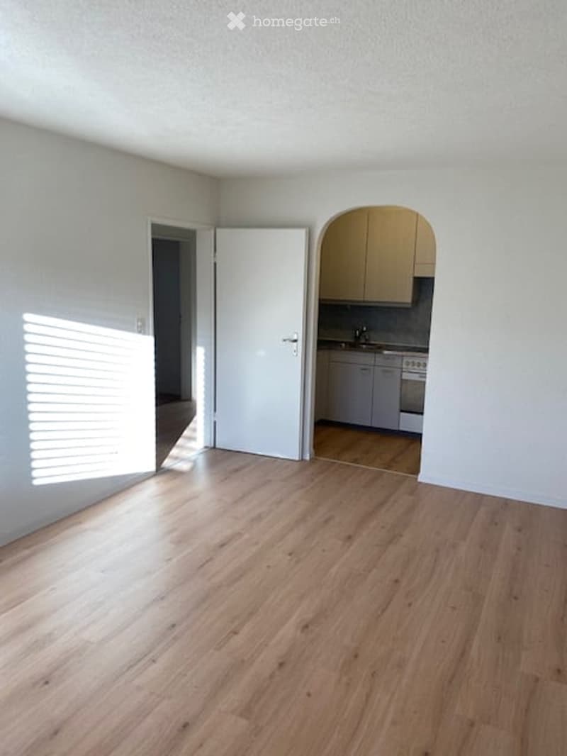 Empty room with light wooden flooring, white walls, and an open doorway leading to a small kitchen area with cabinets and appliances.