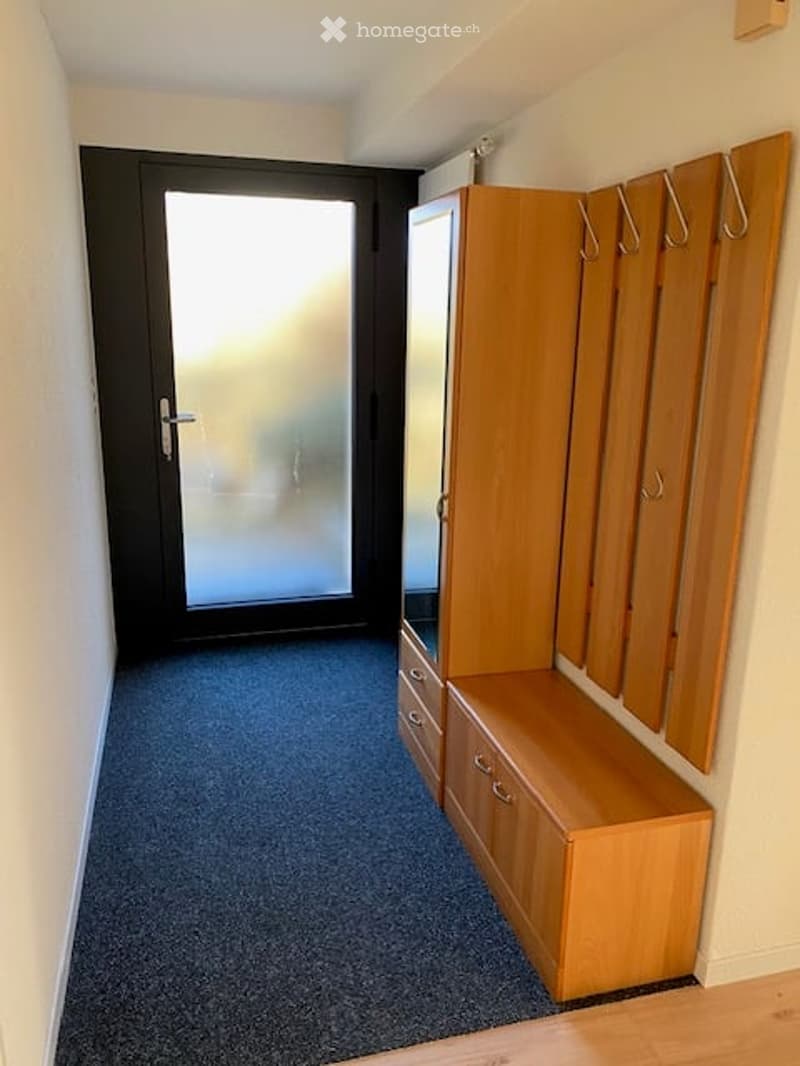 Hallway with a wooden bench and coat rack, leading to a black door with a glass panel.