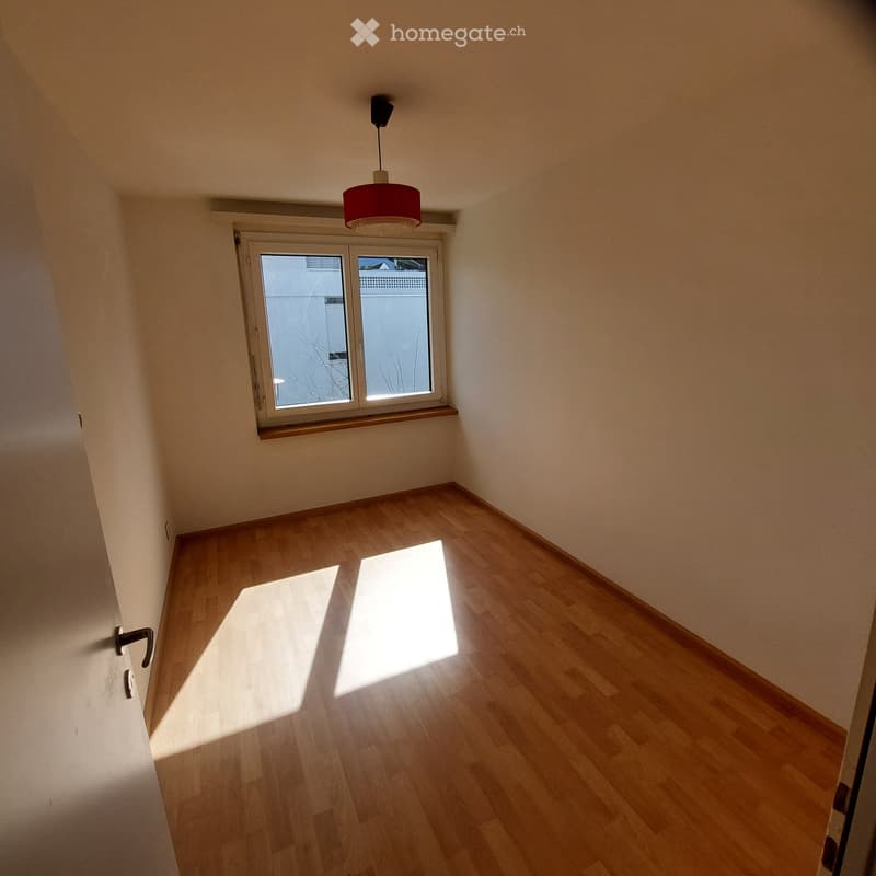Empty room with wooden flooring, white walls, and a single window with blinds. Natural light streams in, casting shadows on the floor. A simple ceiling light fixture is visible.