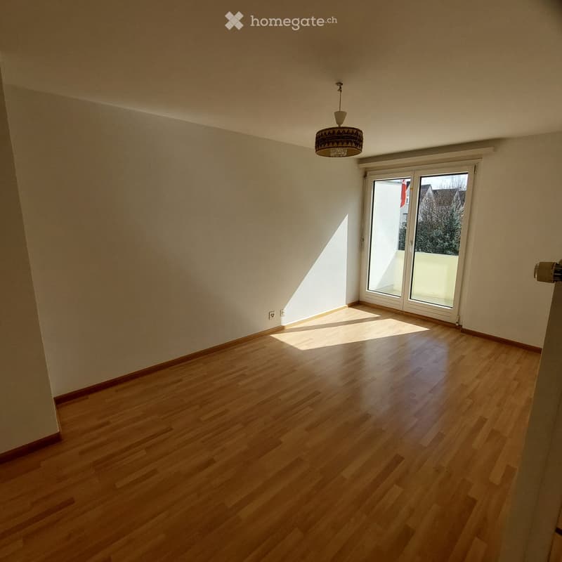 Empty room with wooden flooring, white walls, and a large window with a view of the outside. A hanging light fixture is visible on the ceiling.