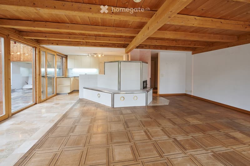 Bright, open room with wooden ceiling beams and herringbone wooden flooring. Features a white kitchen area with cabinets and a countertop, and large glass doors leading to an outdoor space.