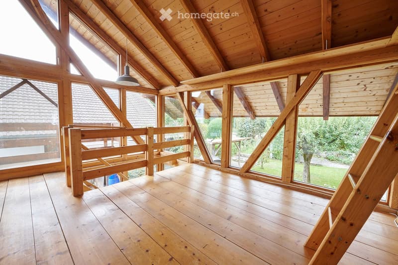 Wooden balcony with slatted railing, exposed wooden beams, and large windows overlooking a garden area.