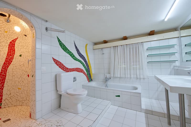 Modern bathroom with white tiles, a bathtub, toilet, sink, and glass shelves. Features a colorful mosaic tile design on one wall and a shower area with a glass curtain.