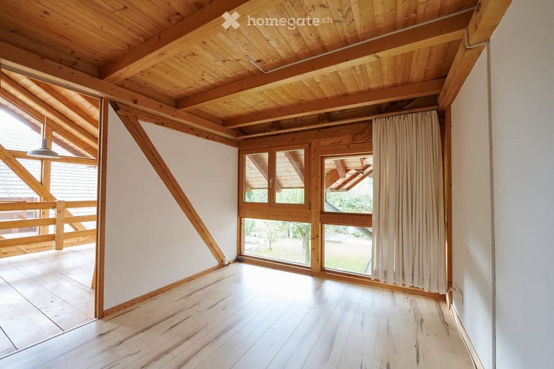 Bright, empty room with wooden floors and exposed wooden beams on the ceiling. Large windows and sliding doors lead to an outdoor area. Minimalist design with white walls and a simple pendant light.