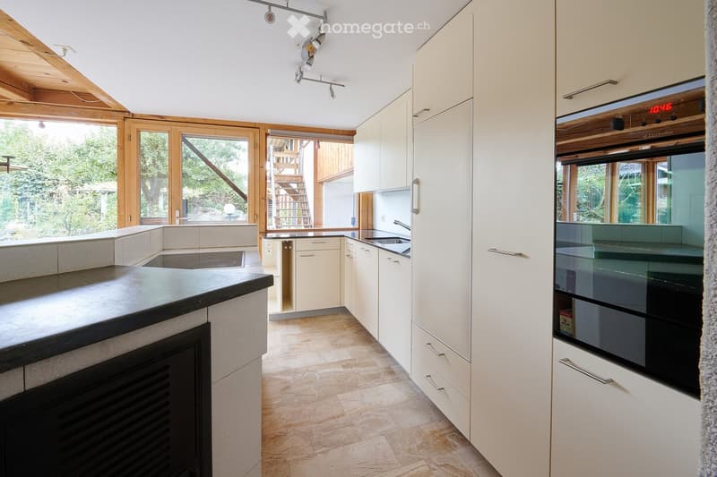 Modern kitchen with white cabinets, black countertop, stainless steel sink, and built-in oven. Large windows provide natural light and access to an outdoor area.