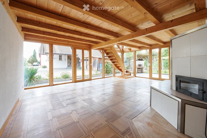 Bright, open room with wooden beams and exposed ceiling, large glass doors leading to outdoor area, wooden staircase, and modern kitchen area with microwave and cabinet.