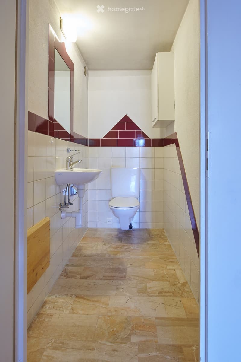 Modern bathroom with white tiles, a sink, toilet, mirror, and a small cabinet. The floor is made of beige stone tiles.