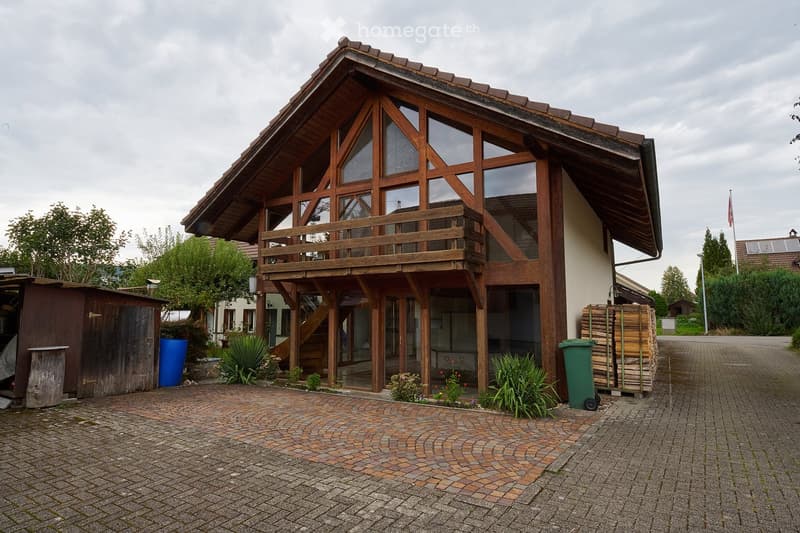 Two-story wooden house with large windows and a balcony, surrounded by greenery and a paved driveway.