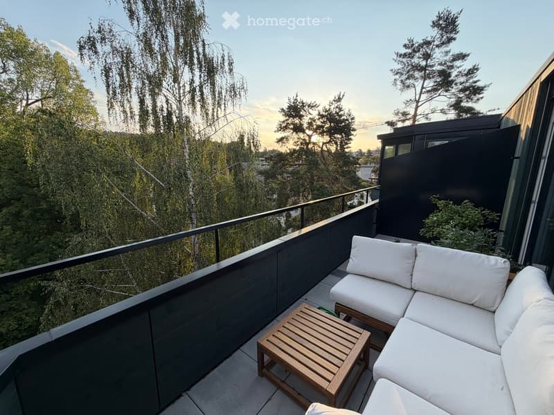 Balcony with a white sectional sofa, wooden coffee table, and lush greenery in the background.