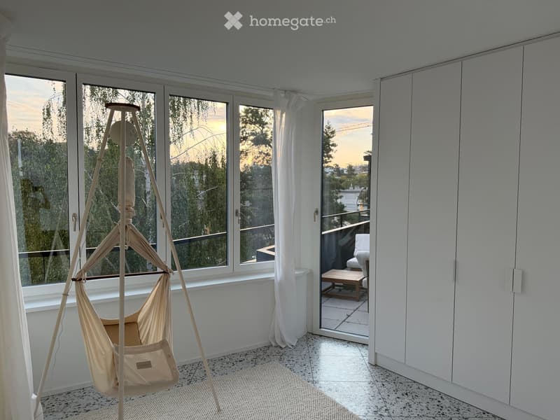 Bright room with large windows and sliding glass doors leading to a balcony. Features a hanging hammock and built-in white cabinetry.