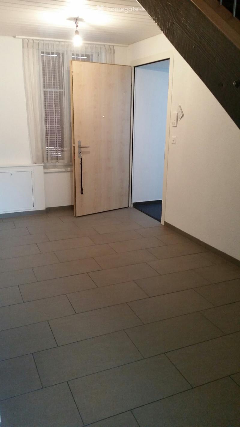 Empty room with tiled floor, white walls, and a wooden door leading to another space. There is a window with blinds and a radiator on the left side.