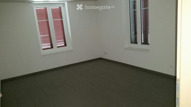 Empty room with light-colored walls, dark tiled floor, and two windows with red blinds. No furniture or equipment present.
