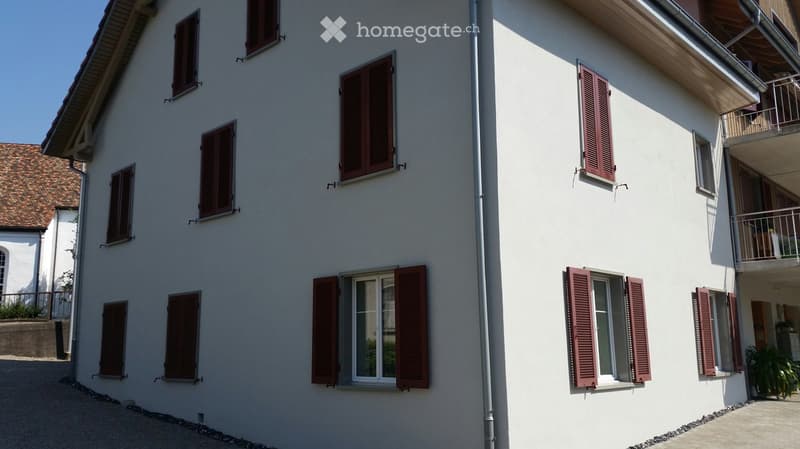 Two-story residential building with white exterior walls, multiple windows with red shutters, and a balcony on the upper floor.