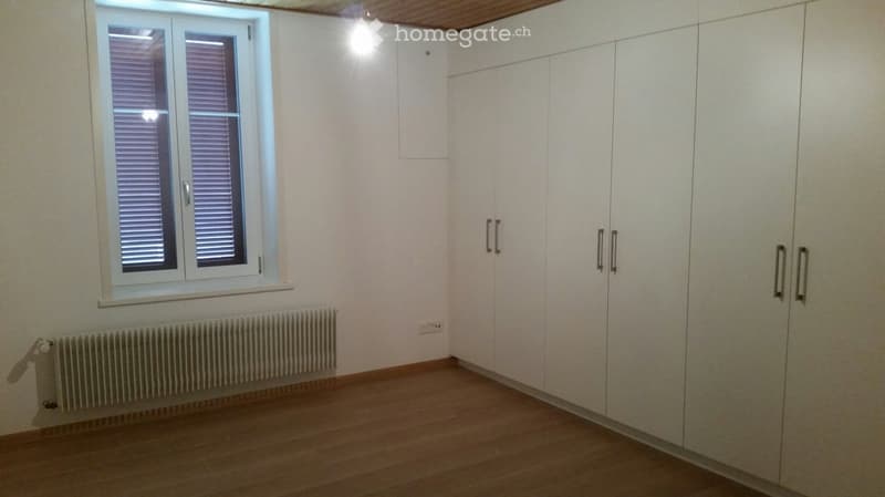 Empty room with light-colored walls, wooden flooring, built-in white wardrobes, a window with blinds, and a radiator below the window.