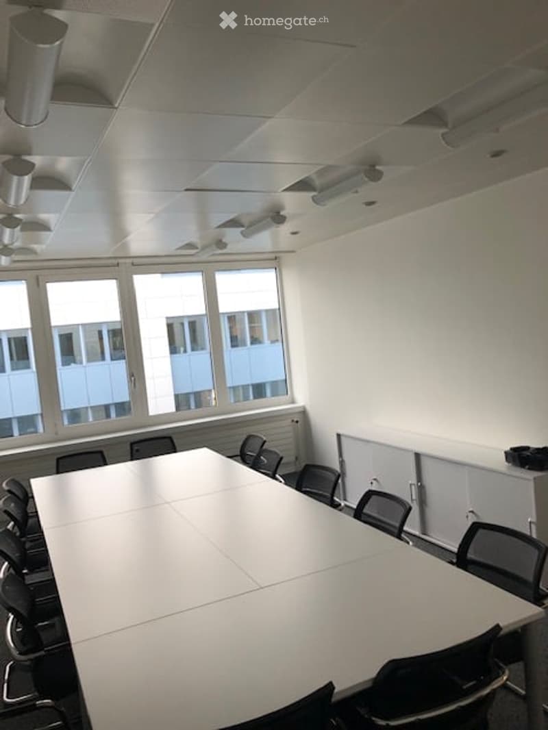 Bright, modern conference room with large windows, a long white table, and multiple black chairs arranged around it. The walls are white, and the ceiling has recessed lighting.