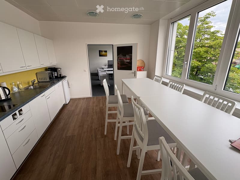 Modern kitchen with white cabinets, a sink, coffee maker, microwave, and a dining table with chairs. The room has wooden flooring and large windows with a view of greenery.