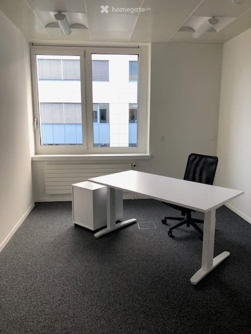 Modern office space with a white desk, black office chair, and a computer tower. Large windows provide natural light, and the room has a grey carpeted floor.