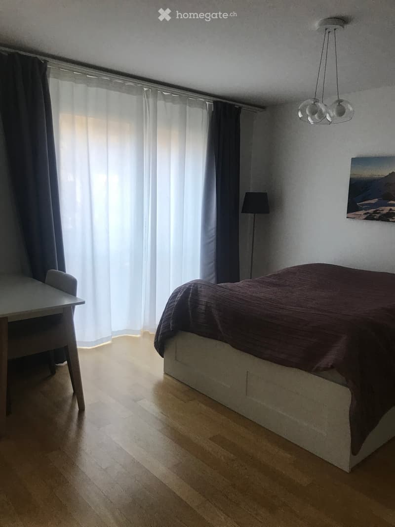 Modern bedroom with wooden flooring, a bed with dark bedding, a small table and chair, a floor lamp, and a picture on the wall.