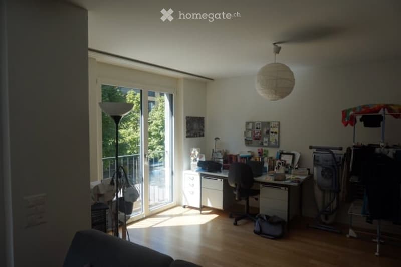 Bright, modern room with large windows and a balcony. Contains a desk with a chair, a lamp, a screen, a table, and a cupboard.