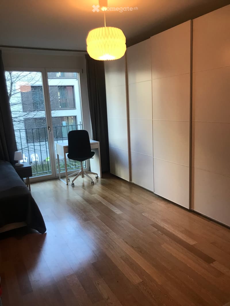 Modern furnished room with wooden flooring, large window with balcony access, a bed, a chair, a table, and a wardrobe.