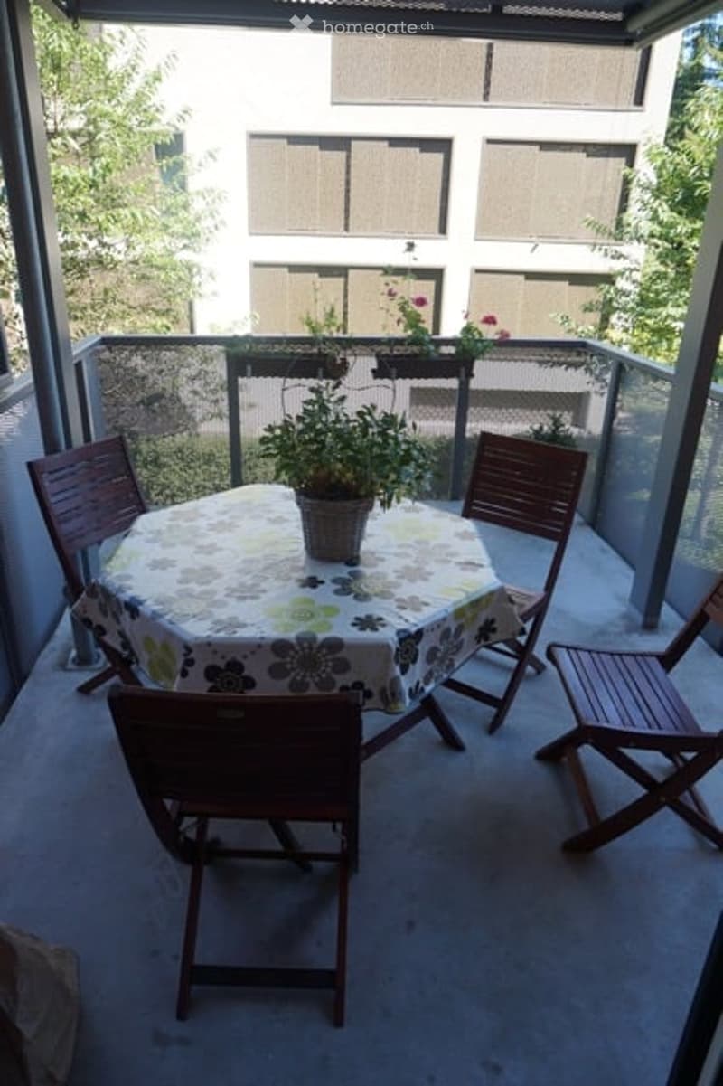 Balcony with a round table covered in a floral tablecloth, surrounded by four wooden chairs, and a potted plant on the table.