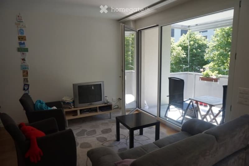 Modern living room with large sliding glass doors leading to a balcony, featuring a grey sofa, coffee table, TV, and chairs.