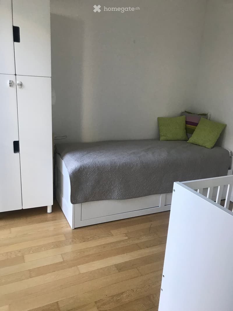 A small bedroom with a white sofa, grey quilted cover, green pillows, and a white wardrobe. Wooden flooring is visible.