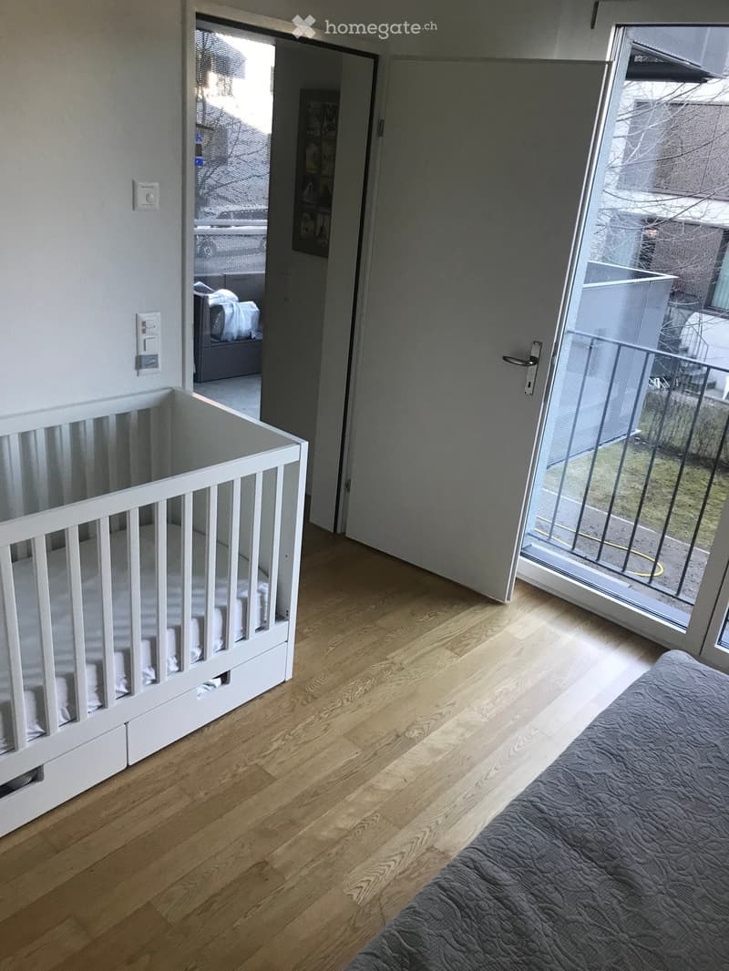 Bright bedroom with wooden flooring, a crib, and a bed, featuring a large window with a view to the balcony.